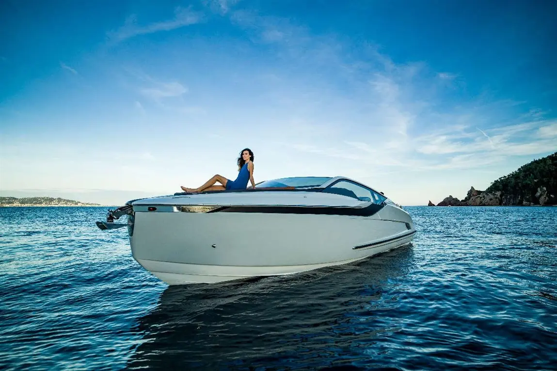 A woman in a blue dress relaxes on the bow of a sleek white yacht amidst calm waters and a clear sky.